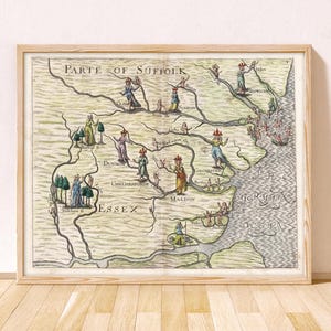 May include: A framed antique-style map titled Parte of Suffolk and Essex, featuring hand-colored illustrations of figures placed across the landscape, winding rivers, and the text The Germain Ocean, all enclosed in a light-toned wooden frame.
