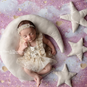 以下が含まれることがあります： 星の装飾が施されたクリーム色の衣装と、お揃いのヘアバンドを身に着けた新生児が、三日月型の枕の上に横たわっています。シーンは、ぬいぐるみ製の星とピンクと紫の背景で飾られています。