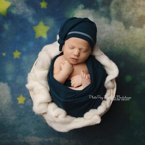 以下が含まれることがあります： 青い星が散りばめられた青と緑の背景に、白いバスケットの中で新生児が眠っています。赤ちゃんはターコイズブルーの毛布にくるまれ、グレーのストライプの入ったターコイズブルーの帽子をかぶっています。