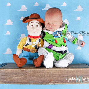 以下が含まれることがあります： 青い雲柄の背景に、緑と白の Buzz Lightyear のコスチュームを着た新生児が、木製棚に座っています。棚には、ウッディのぬいぐるみが置かれています。