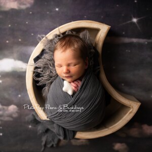 以下が含まれることがあります： 新生児が三日月型の木製小道具の中で眠っています。グレーの包みに包まれ、白い星型の玩具を持っています。背景は濃い青と紫の星空です。