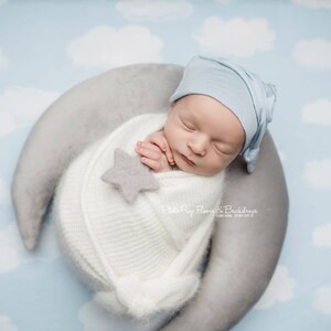以下が含まれることがあります： 新生児が白いニットの包みに包まれ、灰色の三日月型の枕に寄り添って静かに眠っています。赤ちゃんは薄い青色の帽子をかぶっており、包みには小さな灰色の星が置かれています。背景は薄い青色で白い雲があります。