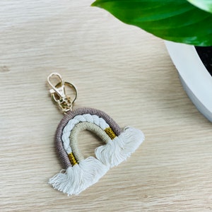 May include: A rainbow-coloured keychain with a gold-coloured metal ring and white tassels. The rainbow is made of braided yarn in shades of beige, white, and light brown.
