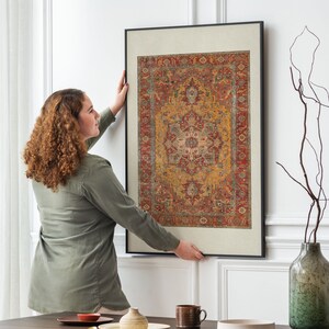 May include: A woman is hanging a framed print of a colourful rug with intricate patterns on a white wall. The rug is predominantly red, orange, and yellow with a central design in shades of pink and white.