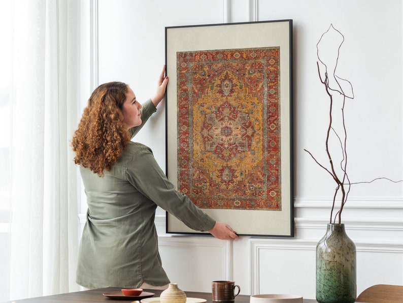 May include: A woman is hanging a framed print of a colourful rug with intricate patterns on a white wall. The rug is predominantly red, orange, and yellow with a central design in shades of pink and white.