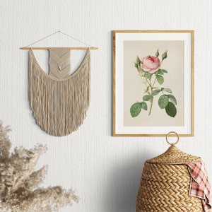 May include: A beige macrame wall hanging with a geometric pattern hangs on a white wall. A framed botanical print of a pink rose with green leaves is on the wall to the right. A wicker basket with a lid sits on the floor below the print.