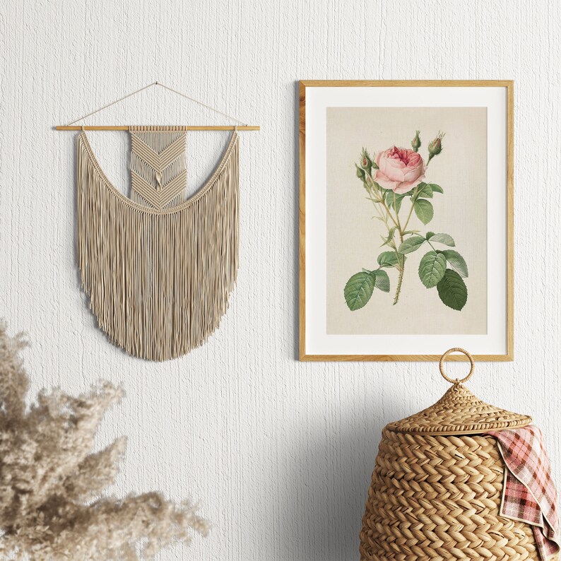 May include: A beige macrame wall hanging with a geometric pattern hangs on a white wall. A framed botanical print of a pink rose with green leaves is on the wall to the right. A wicker basket with a lid sits on the floor below the print.