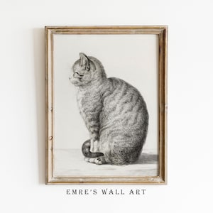 May include: A framed black and white charcoal-style drawing of a tabby cat sitting in profile, facing left. The cat has detailed fur markings and is positioned on a flat surface. The text EMRE'S WALL ART is printed in black at the bottom.