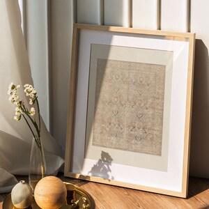 Puede incluir: Un cuadro con marco de madera con un estampado beige con motivos en su interior. El marco est&aacute; sobre un suelo de madera junto a una bandeja dorada con una esfera de madera, un jarr&oacute;n peque&ntilde;o y unas ramitas de flores blancas.