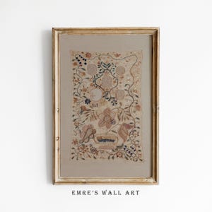 Op de afbeelding: Een ingelijst kunstwerk met een geborduurd ontwerp met bloemen en botanische elementen in tinten beige, bruin en oranje. Het kunstwerk is gevat in een goudkleurige houten lijst. De tekst "EMRE'S WALL ART" is zichtbaar aan de onderkant.