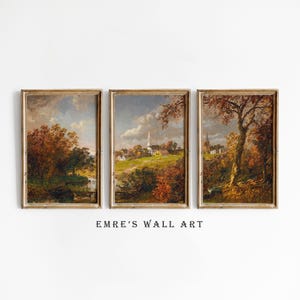 May include: Set of three framed landscape paintings. Each features a detailed autumn scene with trees, a river, and a church. The frames are gold-toned, and the artwork is displayed against a white background. The text "EMRE'S WALL ART" is below the paintings.