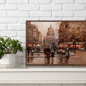 May include: A framed print of a Parisian street scene, featuring a large domed building and figures in period clothing. The artwork is in warm tones, with a focus on architectural details and street activity. A potted plant sits on a white table.