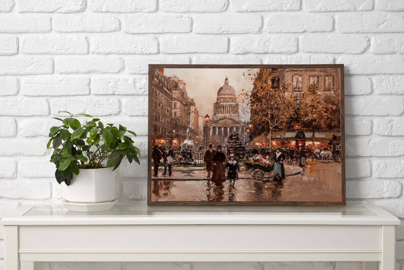May include: A framed print of a Parisian street scene, featuring a large domed building and figures in period clothing. The artwork is in warm tones, with a focus on architectural details and street activity. A potted plant sits on a white table.