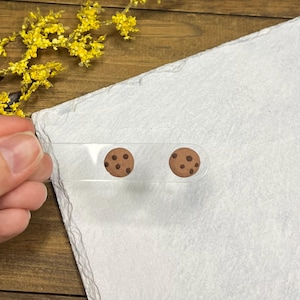 May include: Clear acrylic earrings featuring a chocolate chip cookie design. The earrings are round with brown cookie and chocolate chip details. The earrings are held up against a white background.