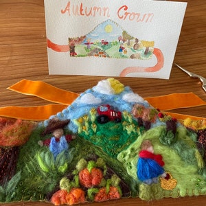 Könnte beinhalten: Eine Filz-Herbstkrone mit einem fantasievollen Landschaftsdesign. Die Krone zeigt einen roten Traktor, einen blauen Himmel mit Wolken und eine Vielzahl von Bäumen, Blumen und Kürbissen. Die Krone ist mit Perlen und Nähten verziert.