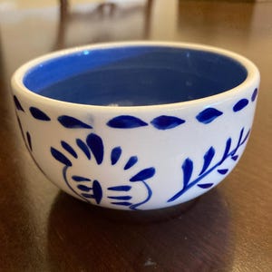 May include: A small ceramic bowl with a white exterior and a deep blue interior. The exterior features hand-painted blue floral and leaf motifs around the sides, with a decorative row of blue dashes encircling the upper rim of the bowl.