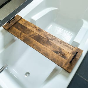 Peut inclure: Un plateau de bain en bois avec une finition marron foncé et des poignées noires. Le plateau rectangulaire s'étend sur la largeur d'une baignoire blanche. Le grain du bois est visible, ajoutant de la texture. Conçu pour les livres ou les boissons.