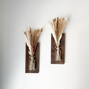 May include: Two wooden wall mounted vases with dried pampas grass in clear glass bottles. The vases are stained a dark brown color.