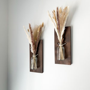 May include: Two wooden wall mounted vases with dried pampas grass in clear glass bottles. The vases are stained a dark brown color.