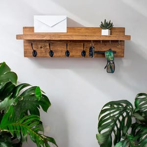 May include: A wooden wall-mounted shelf with five hooks and a small potted plant. The shelf has a natural wood finish and is decorated with a white envelope and sunglasses.