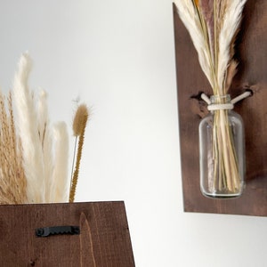May include: Two dark brown wooden wall mounted vases with dried pampas grass and other dried flowers in clear glass bottles. The vases have a black metal hook on the back.