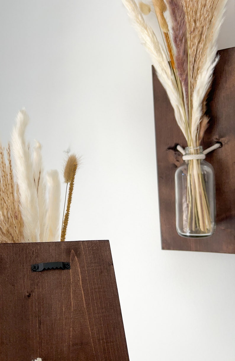 May include: Two dark brown wooden wall mounted vases with dried pampas grass and other dried flowers in clear glass bottles. The vases have a black metal hook on the back.