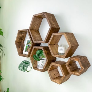 May include: A set of six hexagonal wooden shelves mounted on a white wall. The shelves are arranged in a staggered pattern, with the largest shelf at the top and the smallest shelf at the bottom. The shelves are stained a dark brown color and have a natural wood finish. A small potted plant sits on one of the shelves.