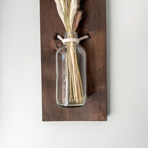 May include: A clear glass vase with dried pampas grass and a white rope tied around the neck of the vase. The vase is mounted on a dark brown wooden board.
