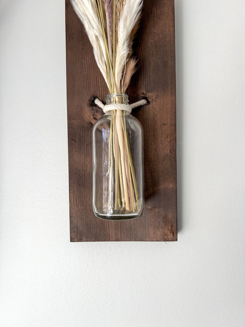 May include: A clear glass vase with dried pampas grass and a white rope tied around the neck of the vase. The vase is mounted on a dark brown wooden board.