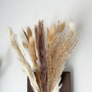 May include: A bouquet of dried pampas grass in shades of brown, beige, and white, arranged in a dark brown wooden vase.