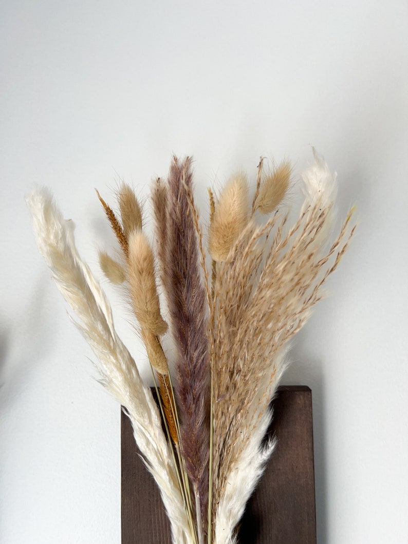 May include: A bouquet of dried pampas grass in shades of brown, beige, and white, arranged in a dark brown wooden vase.