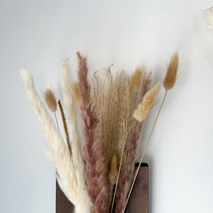 May include: A bouquet of dried pampas grass in shades of white, brown, and pink, arranged in a dark brown wooden vase.