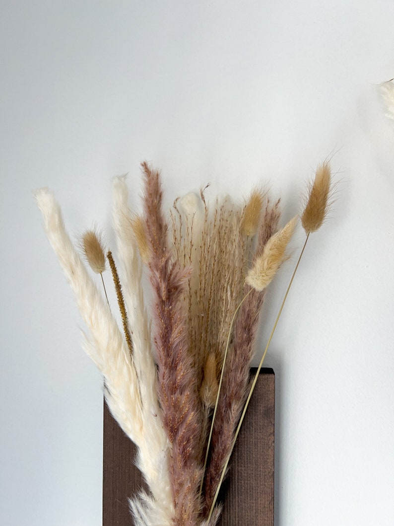 May include: A bouquet of dried pampas grass in shades of white, brown, and pink, arranged in a dark brown wooden vase.
