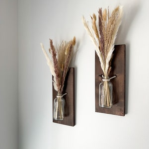 May include: Two brown wooden wall mounted vases with clear glass jars and dried pampas grass in shades of brown, beige, and white.