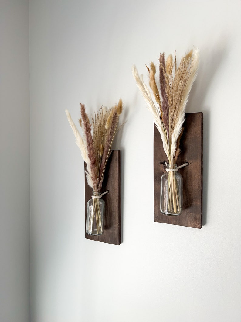May include: Two brown wooden wall mounted vases with clear glass jars and dried pampas grass in shades of brown, beige, and white.