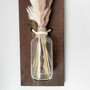 May include: A clear glass vase with a white rope tied around the neck, holding a bouquet of dried pampas grass in shades of white and pink. The vase is mounted on a dark brown wooden wall shelf.