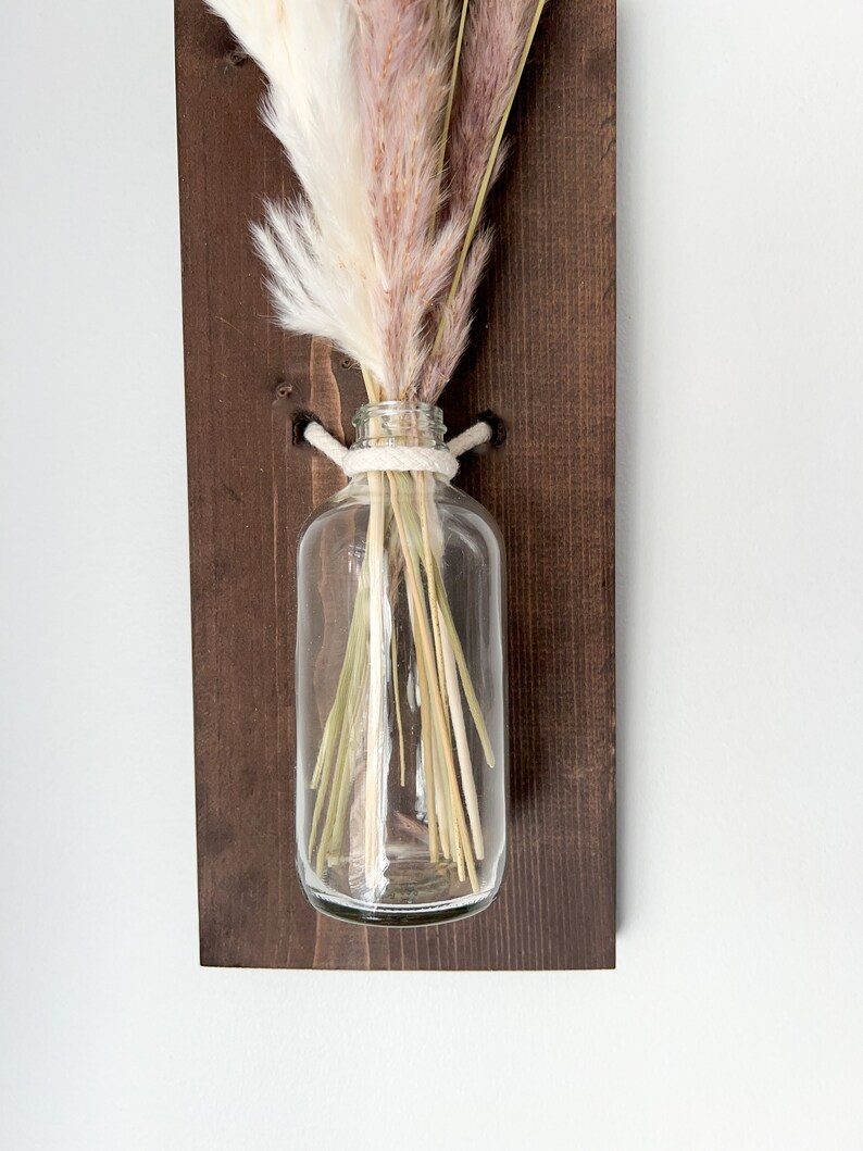 May include: A clear glass vase with a white rope tied around the neck, holding a bouquet of dried pampas grass in shades of white and pink. The vase is mounted on a dark brown wooden wall shelf.