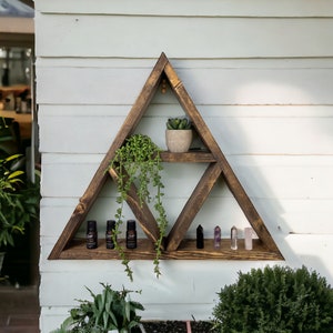 May include: Triangular wooden shelf, stained dark, mounted on a white wall. It displays essential oil bottles, crystals, and potted plants. The shelf's design is geometric, with a triangle shape.