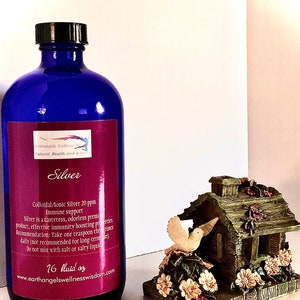 Op de afbeelding: Een blauwe glazen fles met een zilveren etiket waarop "Silver Colloidal/Ionic Silver 20 ppm Immune Support" staat. Het etiket bevat ook gebruiksaanwijzingen en een websiteadres. De fles staat op een wit oppervlak naast een klein, decoratief huis met een vogel op het dak.