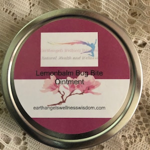 May include: A silver tin with a pink lid and a white label. The label says "Earthangels Wellness Wisdom Natural Health and Wellness Lemonbalm Bug Bite Ointment" and "earthangelswellnesswisdom.com". There is a pink flower illustration on the label.