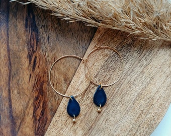 May include: A pair of gold hoop earrings with a teardrop-shaped blue pendant hanging from each hoop. The earrings are on a wooden surface.