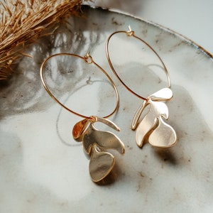 May include: A pair of gold hoop earrings with a leaf-shaped charm dangling from each hoop.