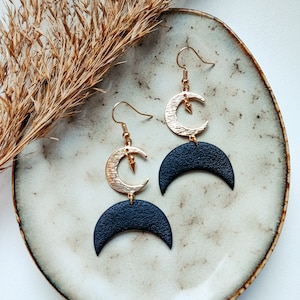 May include: A pair of black and gold crescent moon earrings. The earrings are made of leather and metal. The gold crescent moon has a small gold star in the center.
