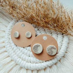 Può includere: Un paio di orecchini a forma di luna bianca e argento con glitter su una carta marrone. La carta è su una superficie di uncinetto bianca.