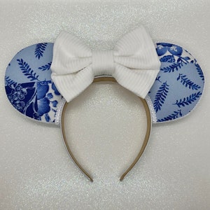 May include: Blue and white floral patterned fabric mouse ears with a white bow. The headband is brown.