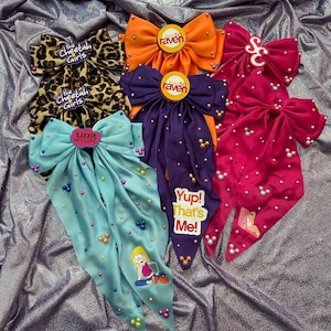 May include: Collection of satin hair bows in various colors, including cheetah print, orange, purple, and pink. Each bow features embellishments and text from popular TV shows. The bows are arranged on a shiny, textured surface.