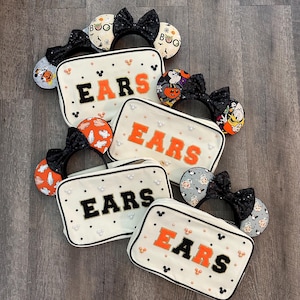 May include: Several off-white rectangular bags with the word "EARS" in orange and black, surrounded by small Mickey Mouse icons and pearls. The bags are paired with various Halloween-themed mouse ear headbands with black sequin bows.