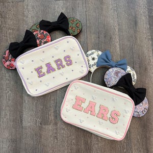 May include: An assortment of decorative headbands and cosmetic bags. The headbands feature various designs, including floral patterns, butterflies, and solid colors, each with a bow. The cosmetic bags are white with the word "EARS" in purple and pink, embellished with pearls.