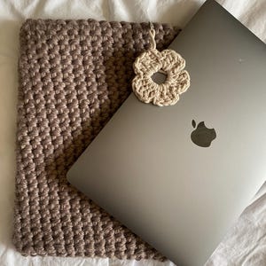 May include: A brown crocheted laptop sleeve with a beige crocheted flower attached. The sleeve is lying on a white surface with a silver laptop inside.