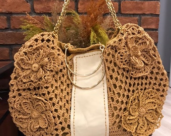 May include: A large, crocheted tote bag with a white leather panel and gold chain straps. The bag is made of brown yarn and features a floral design.